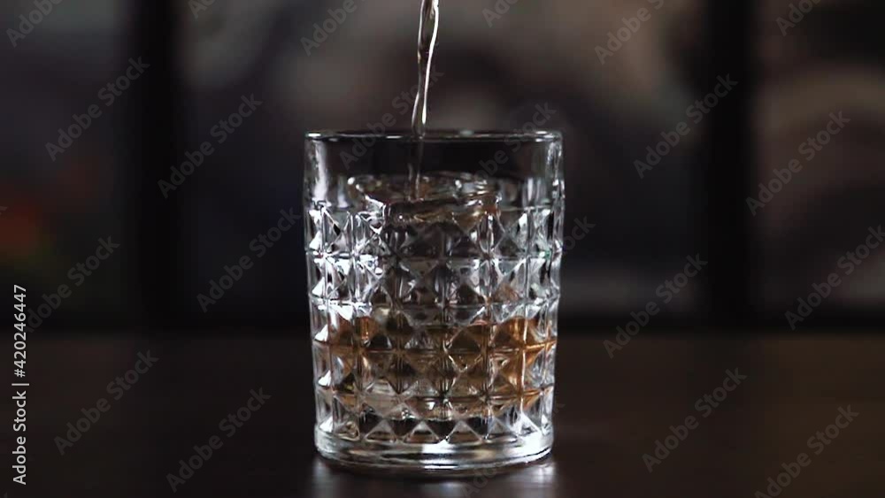 A glass of ice stands on a table with a backlit background, to which is poured an alcoholic cocktail with a thin stream of golden color, which sparkles in the light A glass of ice stands on a table with a backlit background, to which is poured an alcoholic cocktail with a thin stream of golden color, which sparkles in the light