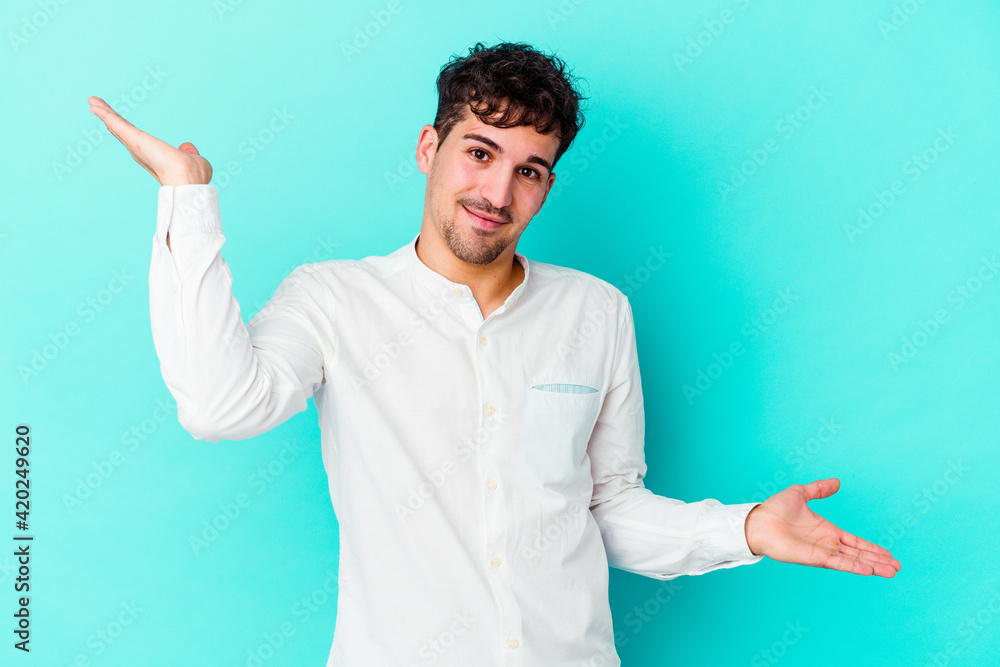 Young caucasian man isolated on blue background makes scale with arms, feels happy and confident. Young caucasian man isolated on blue background makes scale with arms, feels happy and confident.