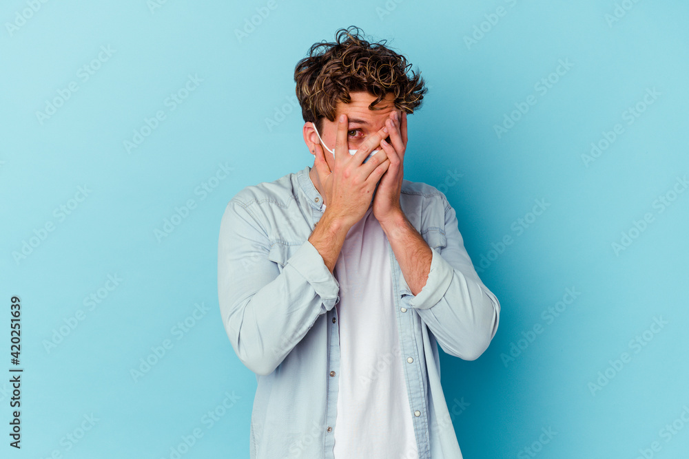 Young caucasian man wearing an antiviral mask isolated on blue background blink through fingers frightened and nervous. Young caucasian man wearing an antiviral mask isolated on blue background blink through fingers frightened and nervous.