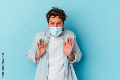 Young caucasian man wearing an antiviral mask isolated on blue background rejecting someone showing a gesture of disgust.