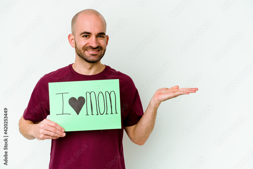 Young caucasian bald man holding a I love mom placard isolated on yellow background showing a copy space on a palm and holding another hand on waist. Young caucasian bald man holding a I love mom placard isolated on yellow background showing a copy space on a palm and holding another hand on waist.