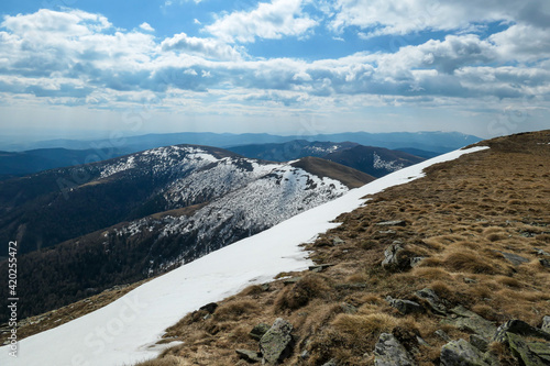 A panoramic view on an Alpine chain in Austria, partially covered with snow. The vast pasture has golden colors. There are other chains in the back. A narrow pathway leading to the top. Overcast photo