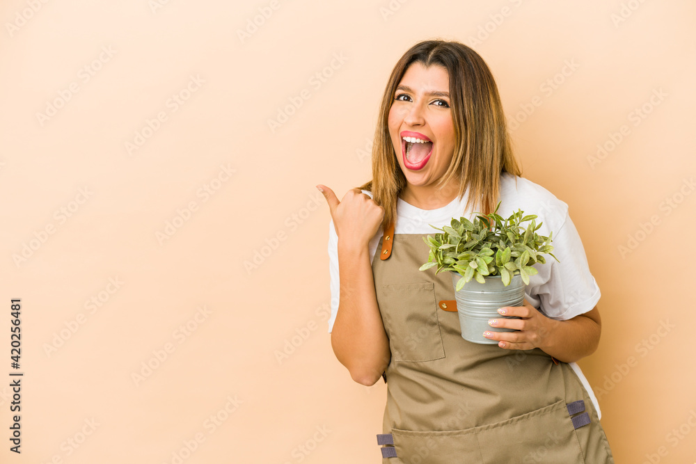 Young indian gardener woman holding a plant isolated points with thumb finger away, laughing and carefree. Young indian gardener woman holding a plant isolated points with thumb finger away, laughing and carefree.