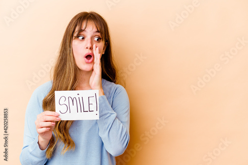 Young caucasian woman holding a Smile placard isolated is saying a secret hot braking news and looking aside