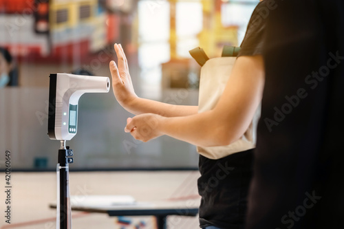 Customer with protective mask raised hand at digital thermometer with temperature scanning machine for fever before entering area. Screening to prevent infected people from entering restricted areas.