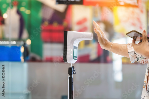 Customer with protective mask raised hand at digital thermometer with temperature scanning machine for fever before entering area. Screening to prevent infected people from entering restricted areas.