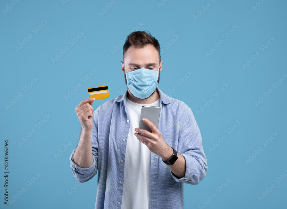 Online shopping during covid. Young guy in face mask purchasing goods on web, holding cellphone and credit card Online shopping during covid. Young guy in face mask purchasing goods on web, holding cellphone and credit card
