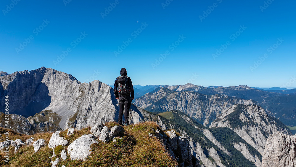 A man with a hiking backpack standing at the edge of a high mountain, Hohe Weichsel in Austria. The man in enjoying the calmness and peace. Lush green pasture around him. Exploration and discovery