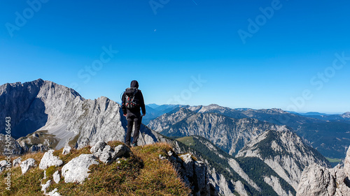 A man with a hiking backpack standing at the edge of a high mountain, Hohe Weichsel in Austria. The man in enjoying the calmness and peace. Lush green pasture around him. Exploration and discovery © Chris