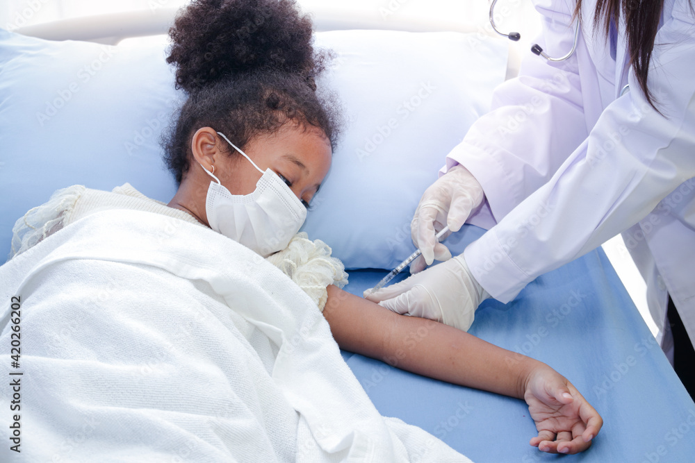 A doctor injects a vaccine in the arm of an African-American girl lying in a hospital bed. To strengthen immunity and protect it from various illnesses. Concept of the vaccine against COVID-19 A doctor injects a vaccine in the arm of an African-American girl lying in a hospital bed. To strengthen immunity and protect it from various illnesses. Concept of the vaccine against COVID-19