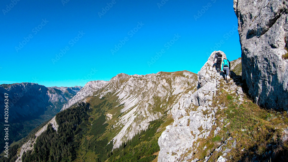 Couple with a hiking backpacks climbing to the of a big boulder on the way to Hohe Weichsel in Austria, with a panoramic view on a vast valley.  Narrow pathway. They are enjoying the view, having fun