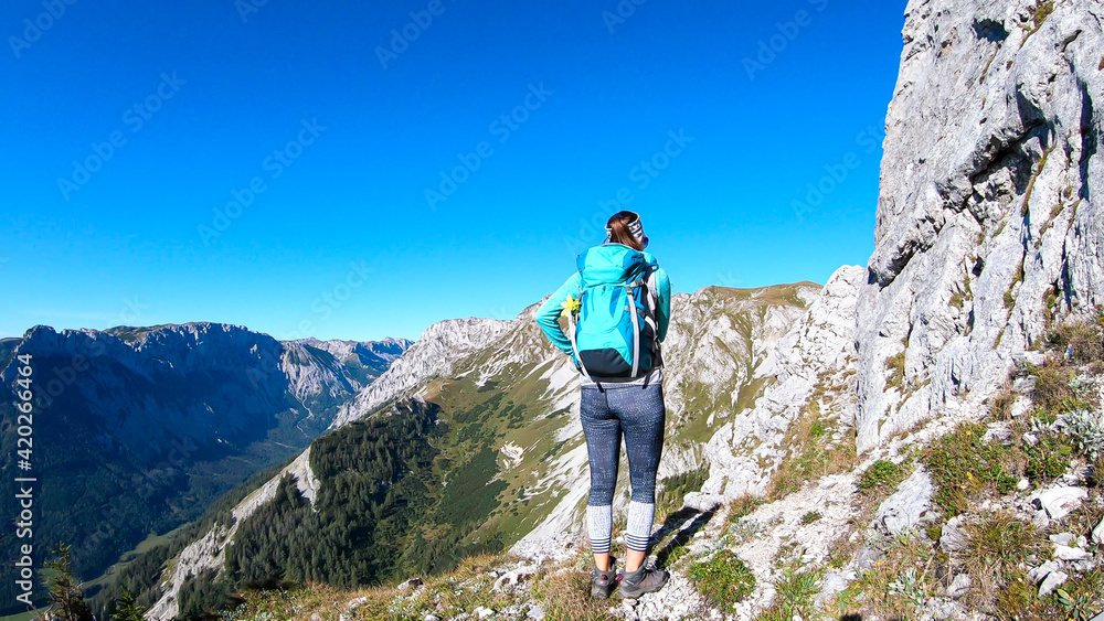 A woman with a hiking backpack climbing to the of a big boulder on the way to Hohe Weichsel in Austria, with a panoramic view on a vast valley.  Narrow pathway. She is enjoying the view. Discovering