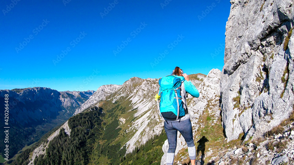 A woman with a hiking backpack climbing to the of a big boulder on the way to Hohe Weichsel in Austria, with a panoramic view on a vast valley.  Narrow pathway. She is enjoying the view. Discovering