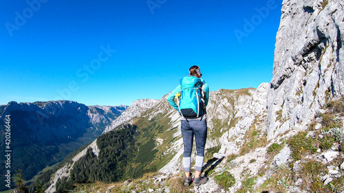 A woman with a hiking backpack climbing to the of a big boulder on the way to Hohe Weichsel in Austria, with a panoramic view on a vast valley. Narrow pathway. She is enjoying the view. Discovering