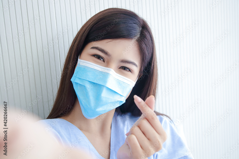 Young female patient wearing a surgical mask Sitting in a hospital bed Hold the phone to chat online with family. Hand made as a mini heart. The concept of health insurance. Young female patient wearing a surgical mask Sitting in a hospital bed Hold the phone to chat online with family. Hand made as a mini heart. The concept of health insurance.