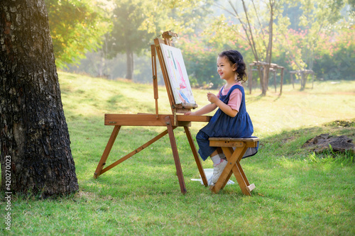A little girl is sitting on the wooden bench and painted on the canvas placed on a drawing stand