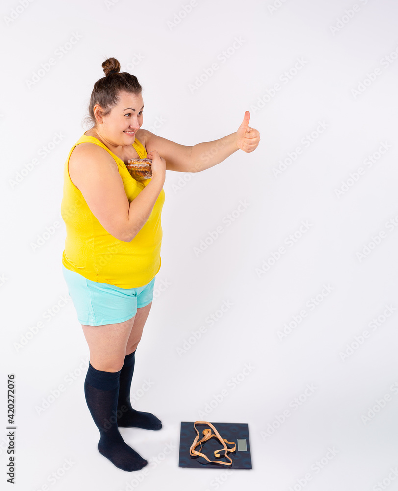 Overweight smiling woman, gesturing thumb up while holding bowl with candy near weighing scale. Woman gourmet measuring body weight holding sweets Overweight smiling woman, gesturing thumb up while holding bowl with candy near weighing scale. Woman gourmet measuring body weight holding sweets
