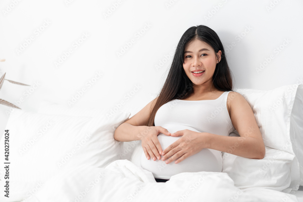 Asian happy pregnant woman is sitting on bed and and touching her belly. pregnancy, motherhood, people and expectation concept Asian happy pregnant woman is sitting on bed and and touching her belly. pregnancy, motherhood, people and expectation concept