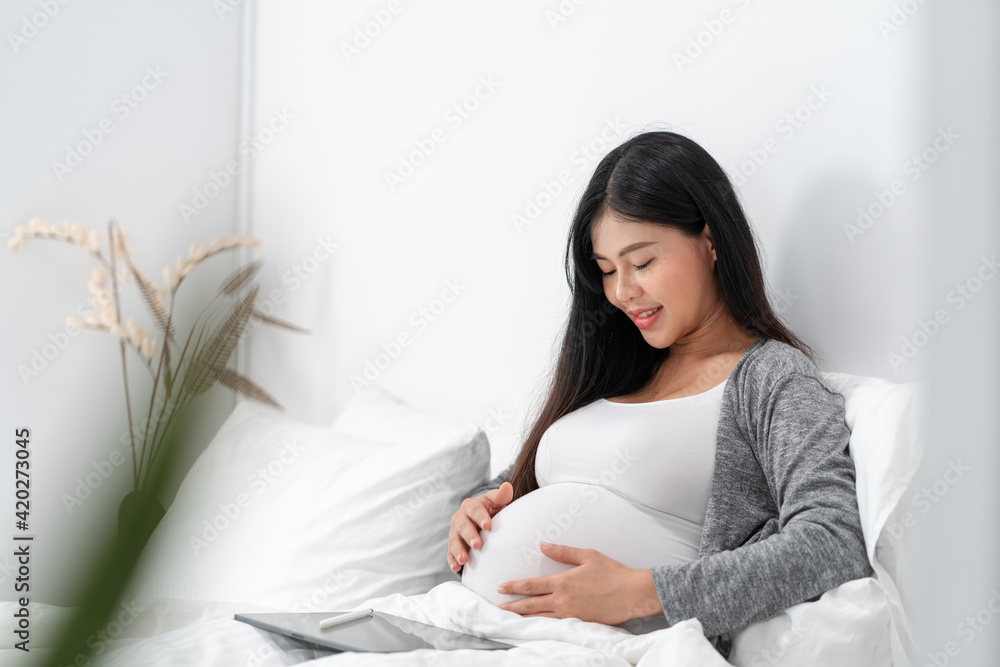 Asian happy pregnant woman is sitting and reading a book on bed and and touching her belly. pregnancy, motherhood, people and expectation concept Asian happy pregnant woman is sitting and reading a book on bed and and touching her belly. pregnancy, motherhood, people and expectation concept