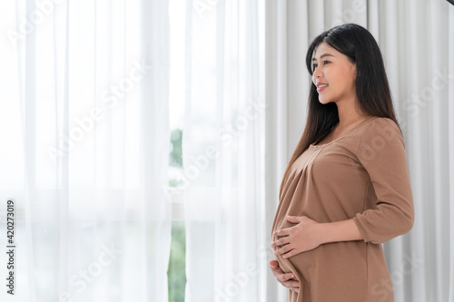 Asian happy pregnant woman is standing beside windows and touching her belly. pregnancy, motherhood, people and expectation concept