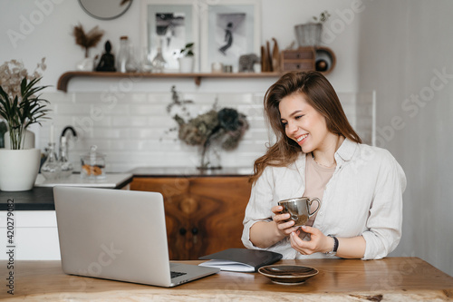Happy attractive young woman sitting in her kitchen at the counter with a laptop smiling at the camera