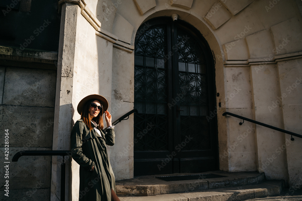 Outdoor fashion portrait of elegant, luxury woman wearing beige hat, sunglasses, trendy white shirt, in a green trench coat, walking in street. Copy, empty space for text