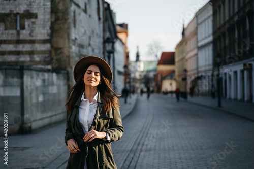Outdoor fashion portrait of elegant, luxury woman wearing beige hat, sunglasses, trendy white shirt, in a green trench coat, walking in street. Copy, empty space for text © Oleksandr