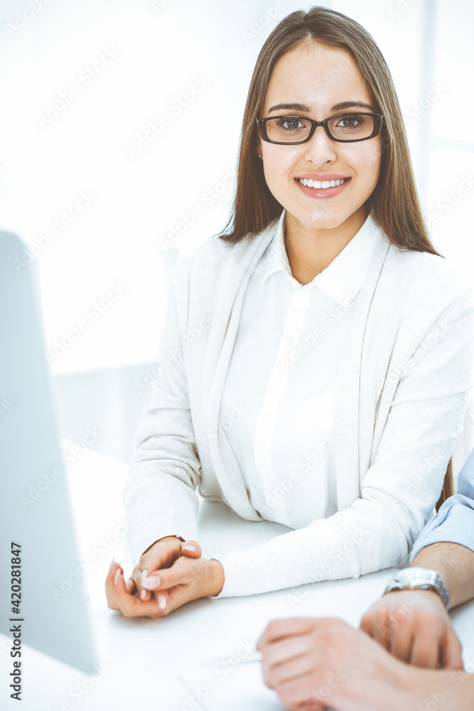 Young cheerful woman sitting at the desk with computer and looking at camera in white colored office. Looks like student girl or business lady. Headshot Young cheerful woman sitting at the desk with computer and looking at camera in white colored office. Looks like student girl or business lady. Headshot