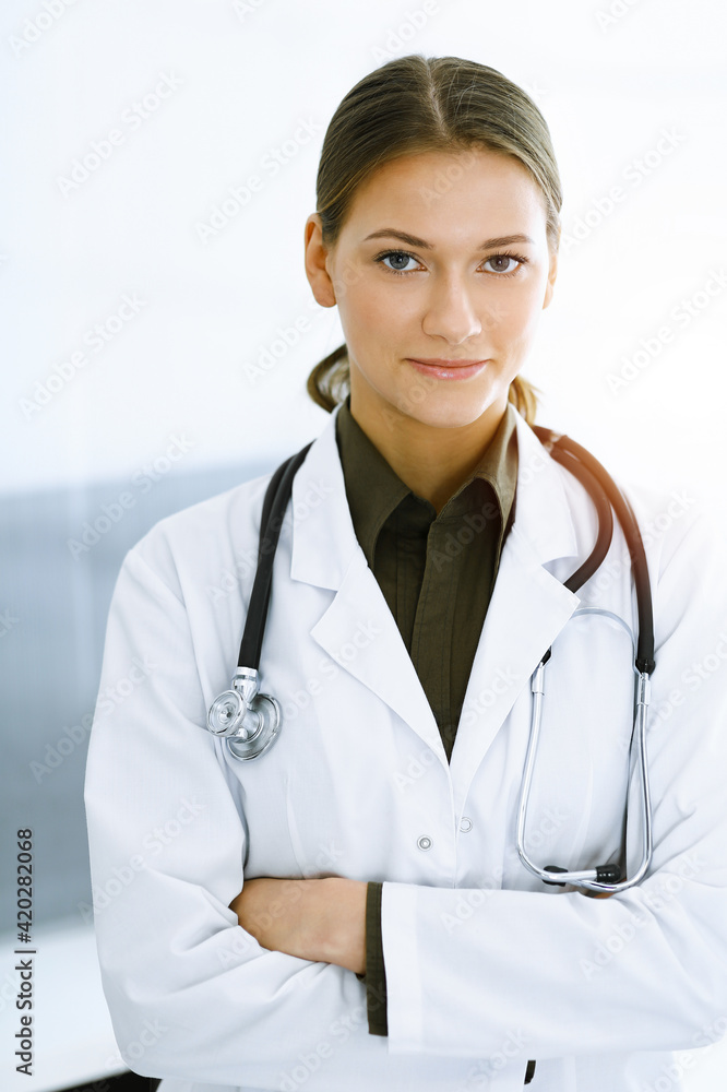 Woman-doctor standing with arms crossed and looking at camera in sunny clinic. Medicine and healthcare Woman-doctor standing with arms crossed and looking at camera in sunny clinic. Medicine and healthcare