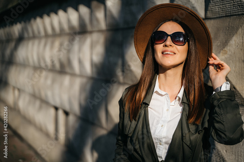 Outdoor fashion portrait of elegant, luxury woman wearing beige hat, sunglasses, trendy white shirt, in a green trench coat, walking in street. Copy, empty space for text © Oleksandr