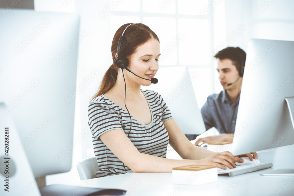Casual dressed young woman using headset and computer while talking with customers online. Group of operators at work. Call center, business concept Casual dressed young woman using headset and computer while talking with customers online. Group of operators at work. Call center, business concept