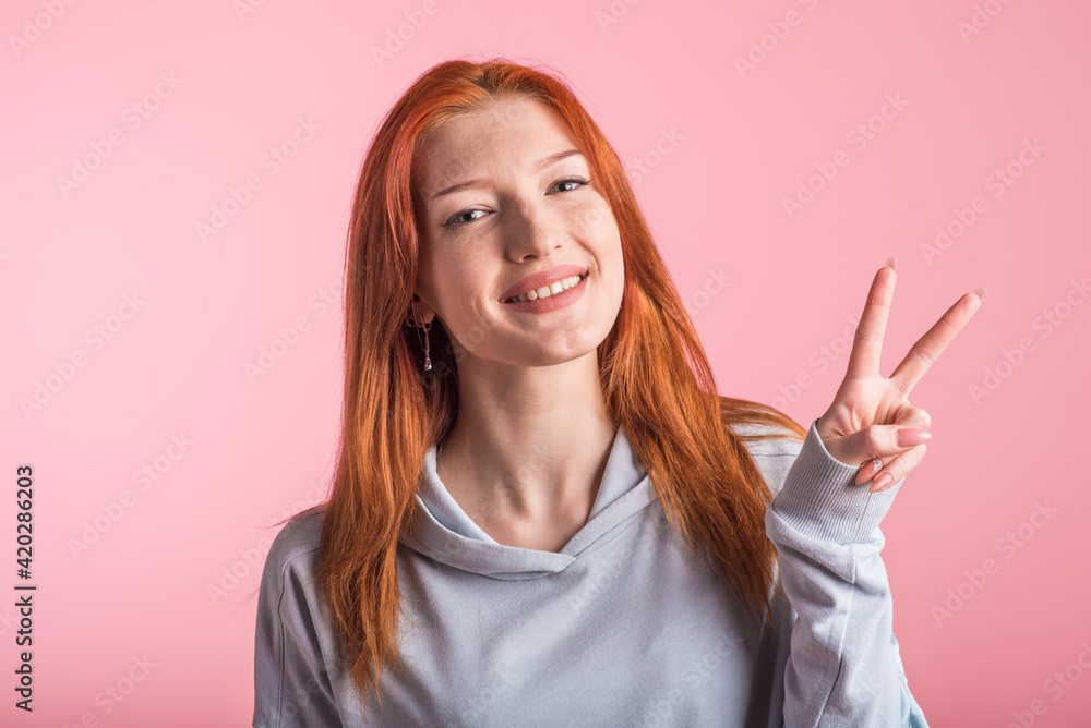 Young redhead girl showing peace gesture in studio on pink background Young redhead girl showing peace gesture in studio on pink background