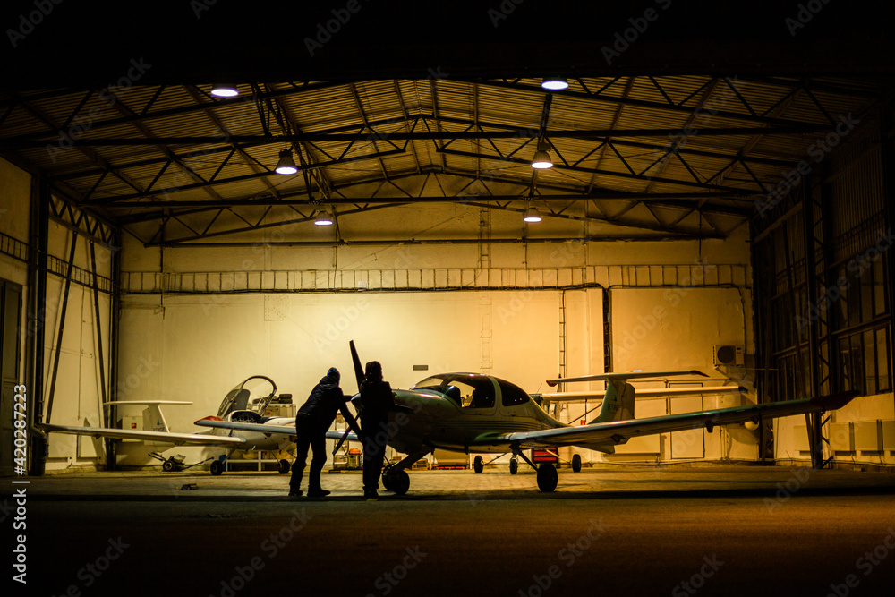 two men parking small jet in a hangar at night - obrazy, fototapety, plakaty 
