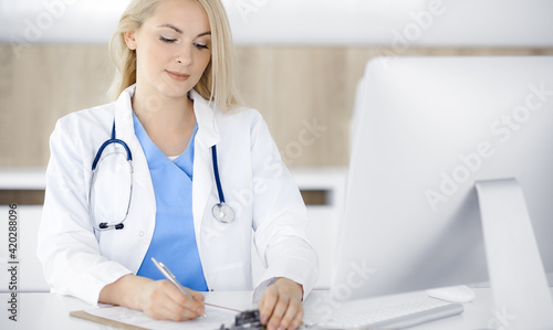 Woman-doctor at work while sitting at the desk in hospital or clinic. Blonde cheerful physician filling up medication history record form