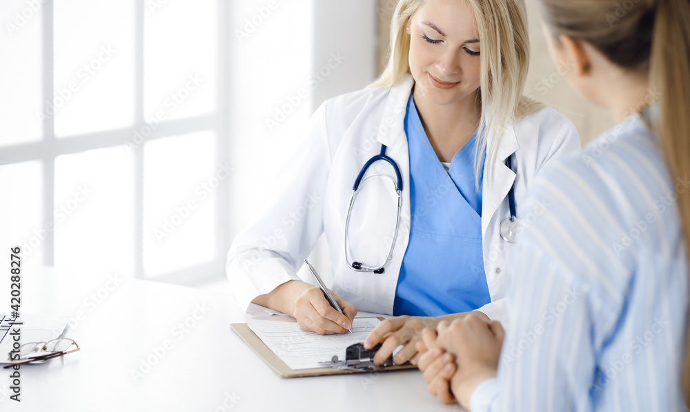 Woman-doctor and patient sitting and talking in hospital or clinic. Blonde therapist is cheerfully smiling. Medicine concept Woman-doctor and patient sitting and talking in hospital or clinic. Blonde therapist is cheerfully smiling. Medicine concept