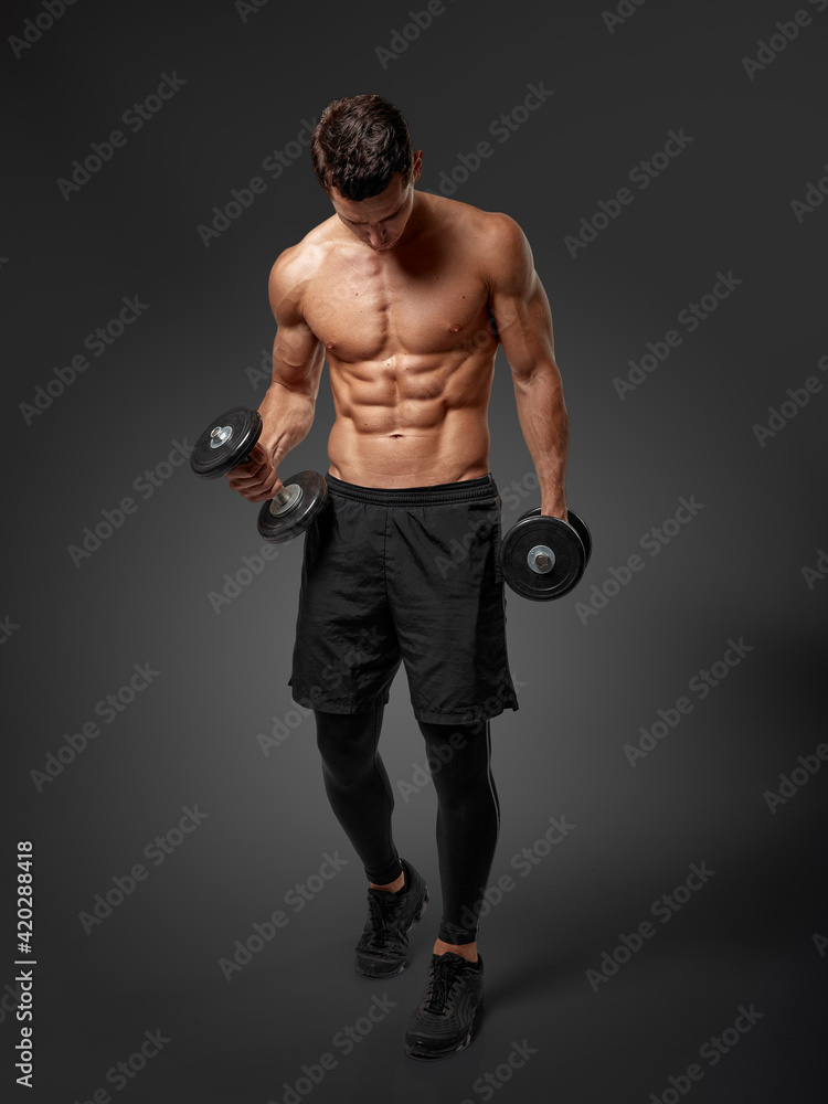 Full length image of a confident man shirtless torso portrait training with dumb-bell, looking down, grey background.