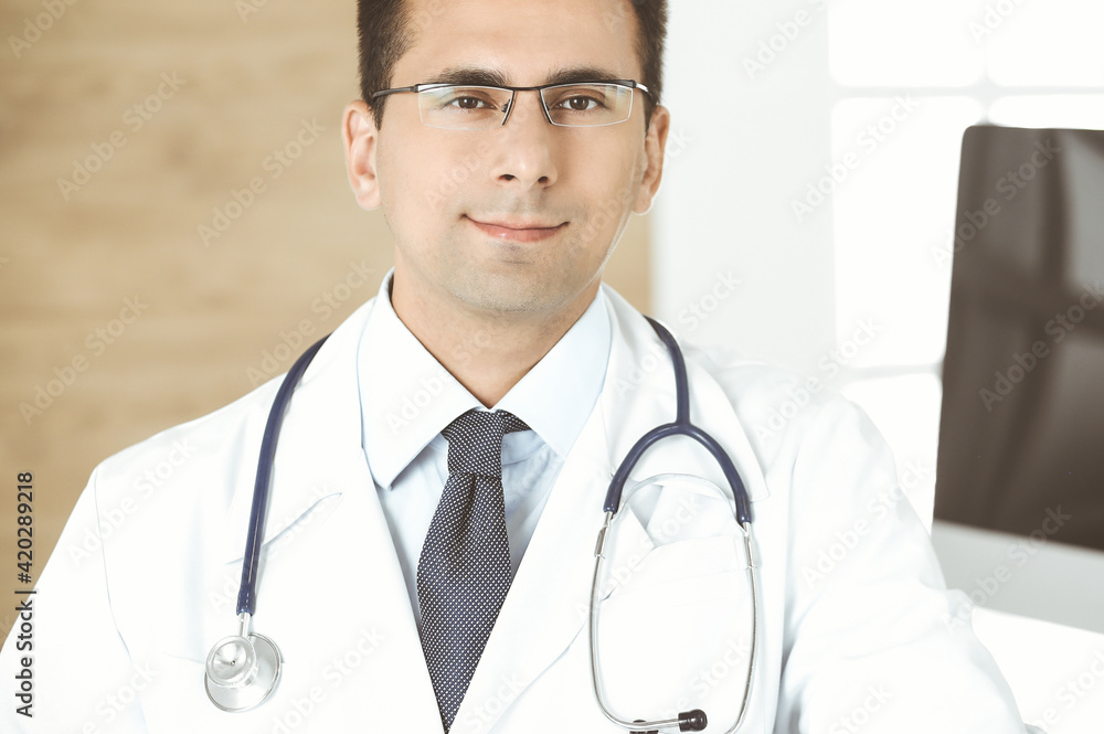Man-doctor sitting at the desk at his working place and smiling at camera. Perfect medical service in clinic Man-doctor sitting at the desk at his working place and smiling at camera. Perfect medical service in clinic