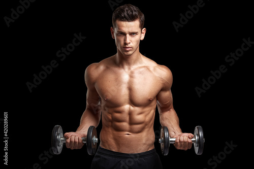 Front image of a fit, strong athletic young man with bare torso, training with dumb-bell, isolated black background.