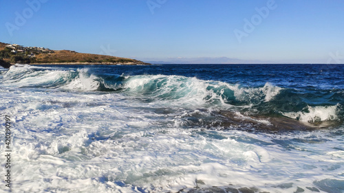 Sunny clear stormy wave in blue sea hitting rocky shore. Summer sea with white waves on Aegean sea in Athens, Greece