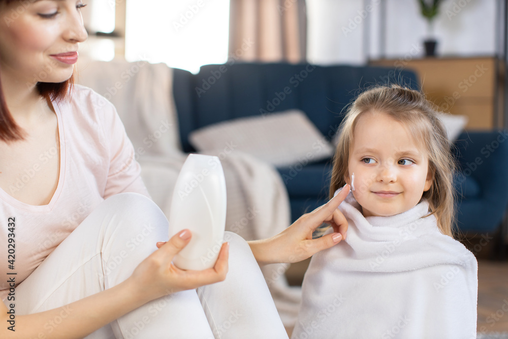 Close up of young smiling mother applying moisturizing lotion or cream on her cute daughter s face after bathing at home. Children skin care and hygiene concept Close up of young smiling mother applying moisturizing lotion or cream on her cute daughter s face after bathing at home. Children skin care and hygiene concept