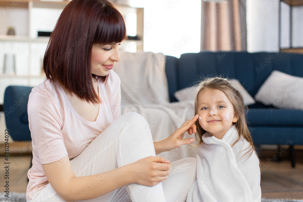 Young attractive mother taking care of her little daughter in a towel after bath, applying body lotion or cream on baby cheeks. Skin home care, children hygiene and healthy lifestyle Young attractive mother taking care of her little daughter in a towel after bath, applying body lotion or cream on baby cheeks. Skin home care, children hygiene and healthy lifestyle