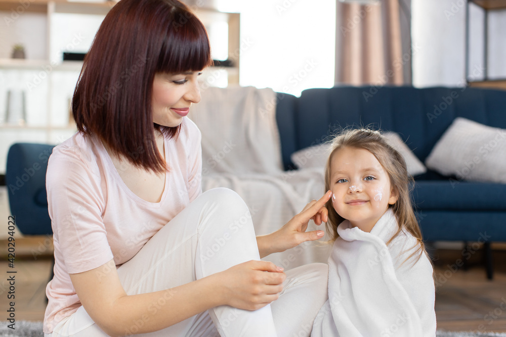 Young attractive mother taking care of her little daughter in a towel after bath, applying body lotion or cream on baby cheeks. Skin home care, children hygiene and healthy lifestyle