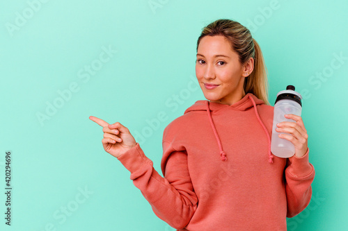 Young caucasian sport woman drinking water isolated on blue backgroundsmiling and pointing aside, showing something at blank space. photo