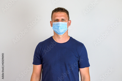 Portrait of young man. He is wearing blue protective face mask and shirt with short sleeves. White background. Preventive measures.