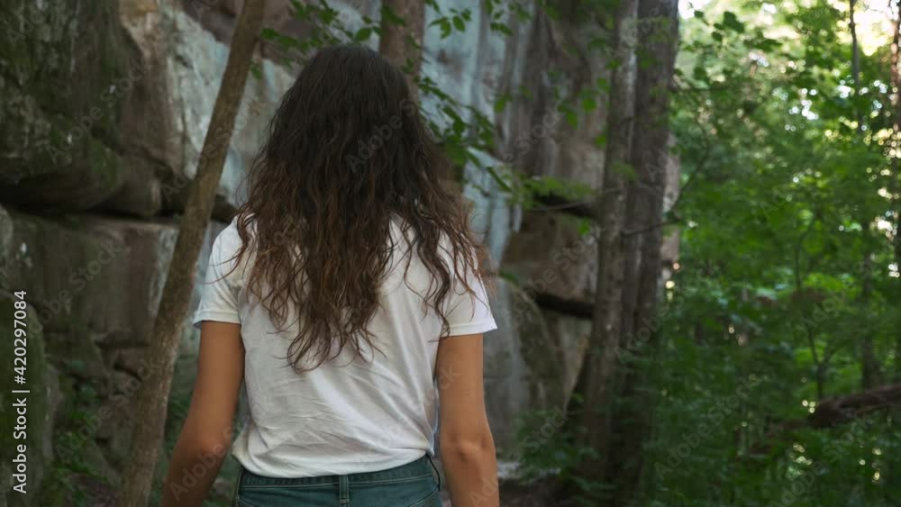 Young hipster woman out hiking along a sunny Tennessee trail with tall trees and rock wall cliff, slow motion cinematic
