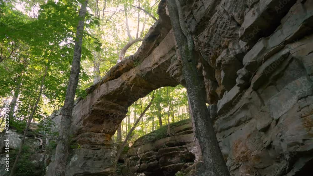 Huge natural rock bridge in a beautiful forest, sunrays, camera pushing forward and looking up in slow motion, young woman exploring and hiking across Huge natural rock bridge in a beautiful forest, sunrays, camera pushing forward and looking up in slow motion, young woman exploring and hiking across