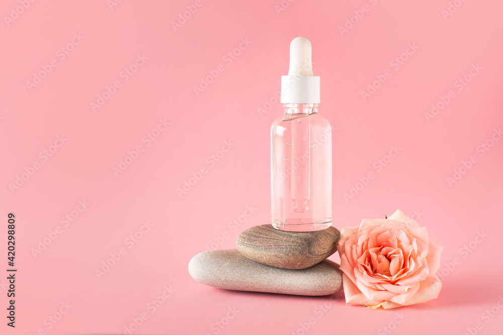 Bottle of facial serum on stone with rose flower on pink background