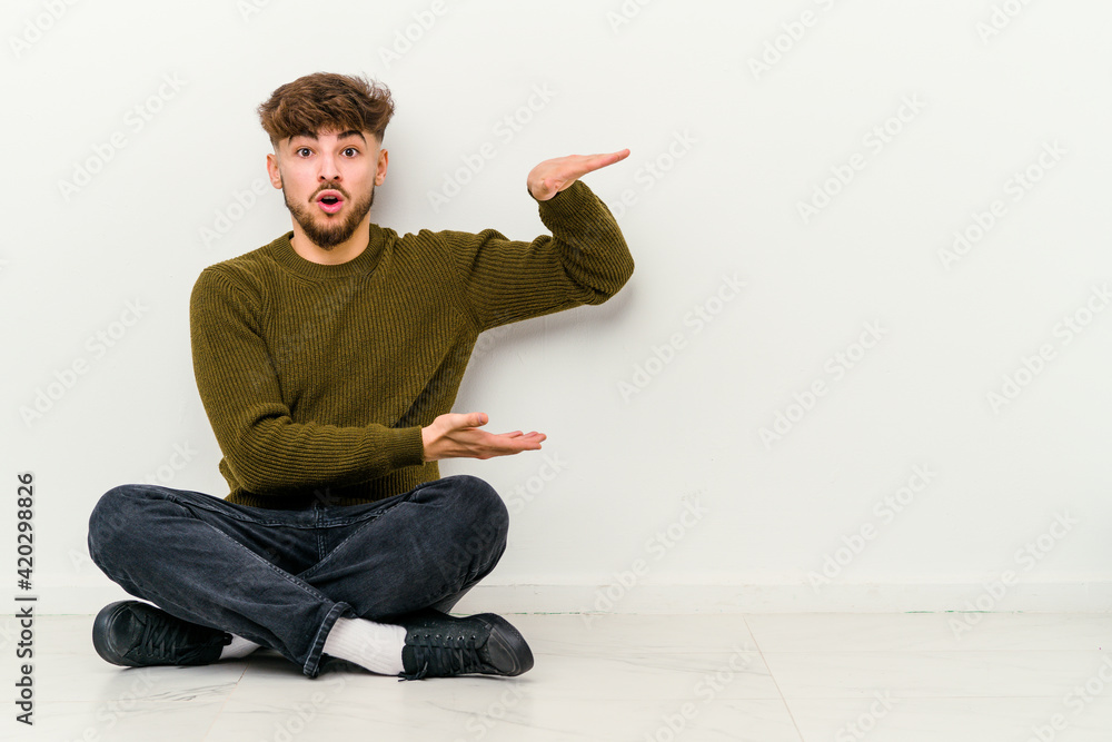 Young Moroccan man sitting on the floor isolated on white background shocked and amazed holding a copy space between hands. Young Moroccan man sitting on the floor isolated on white background shocked and amazed holding a copy space between hands.