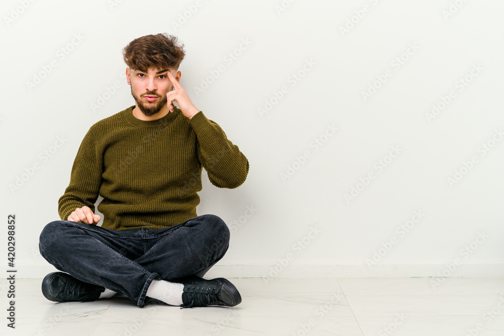 Young Moroccan man sitting on the floor isolated on white background pointing temple with finger, thinking, focused on a task. Young Moroccan man sitting on the floor isolated on white background pointing temple with finger, thinking, focused on a task.