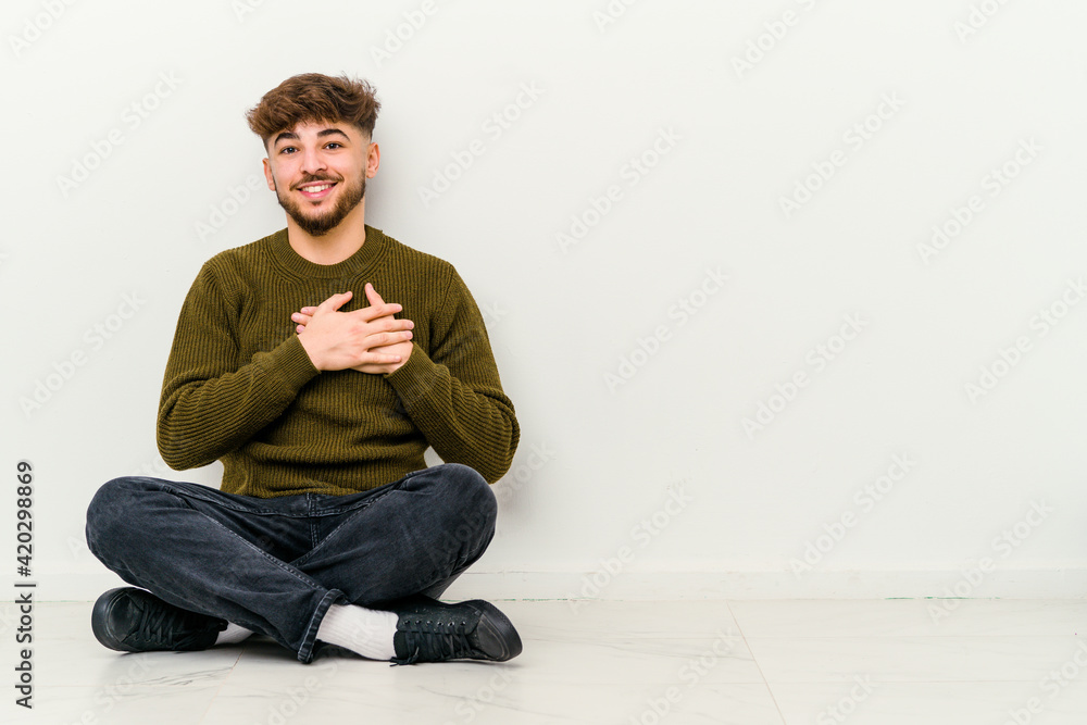 Young Moroccan man sitting on the floor isolated on white background has friendly expression, pressing palm to chest. Love concept.